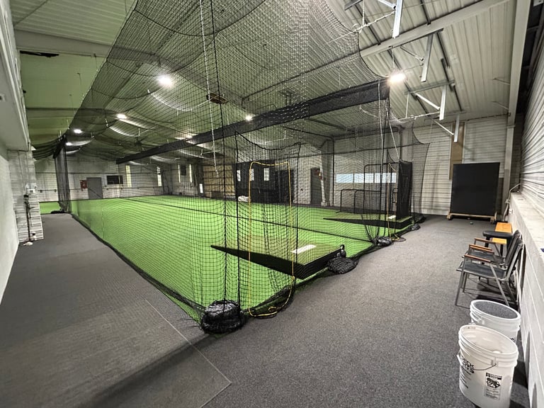 Indoor sports facility with bright green turf field enclosed by black netting, industrial ceiling lights, and equipment storage along the walls