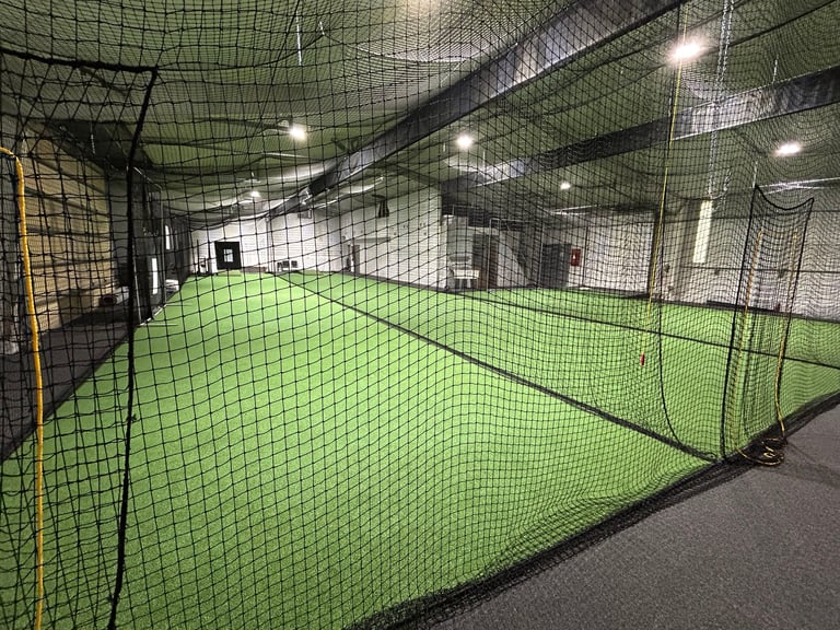 Indoor tennis court with bright green playing surface enclosed by black netting and metal framework